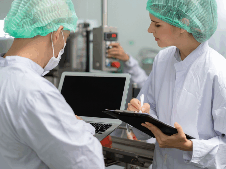 Profissionais de laboratório em ambiente controlado analisando dados em laptop e registrando informações em prancheta, simbolizando rastreabilidade, controle de qualidade e conformidade com as Boas Práticas de Laboratório (BPL).