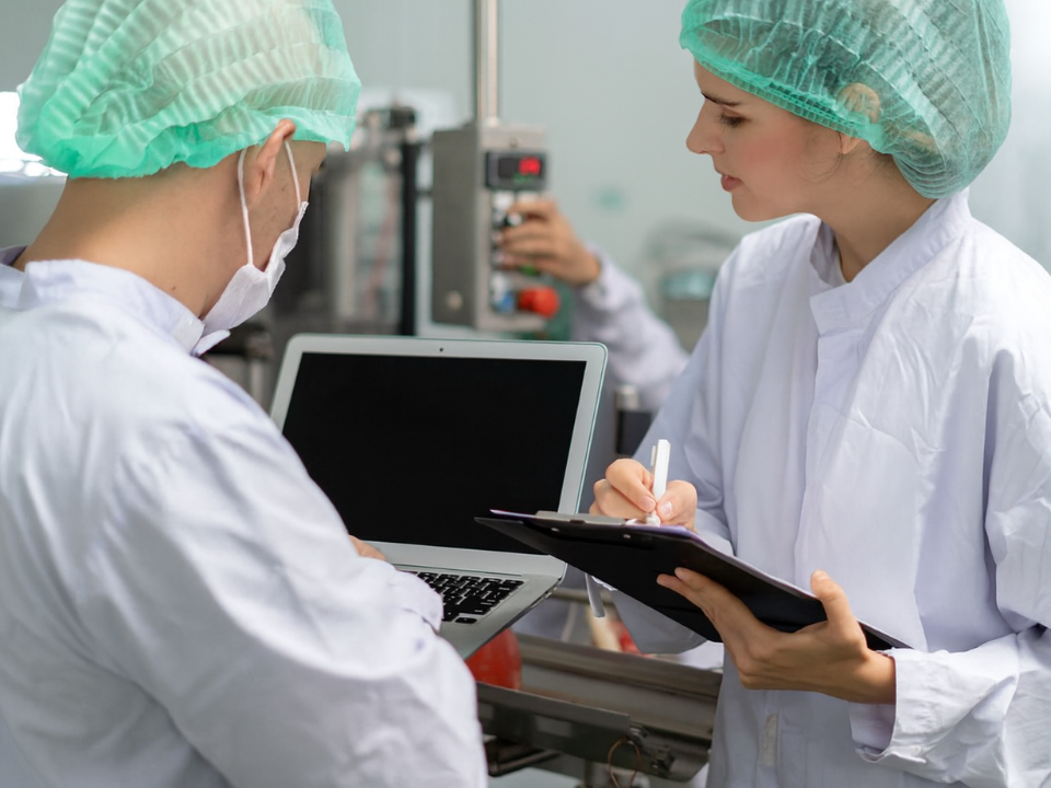 Profissionais de laboratório em ambiente controlado analisando dados em laptop e registrando informações em prancheta, simbolizando rastreabilidade, controle de qualidade e conformidade com as Boas Práticas de Laboratório (BPL).