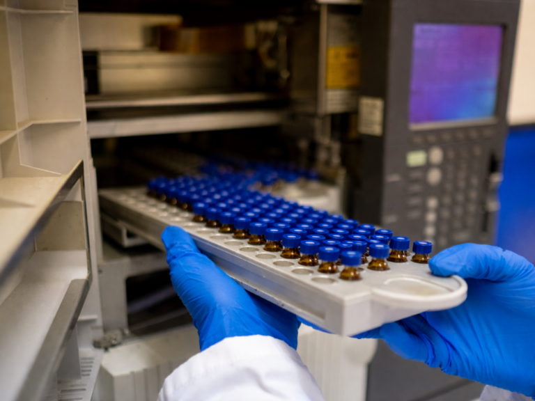 Analista em laboratório de controle de qualidade posiciona bandeja de amostras com frascos em autosampler de sistema HPLC, evidenciando rotina de cromatografia líquida e monitoramento do desempenho instrumental.