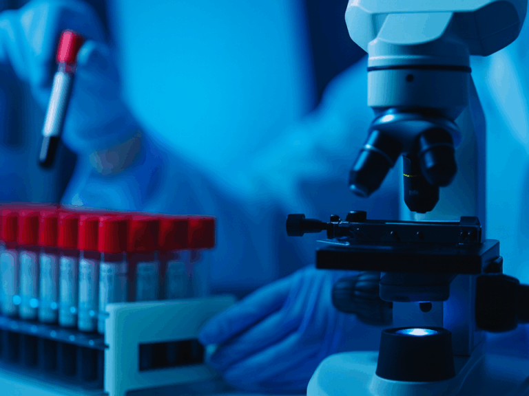 Microscópio em primeiro plano e profissional de laboratório manuseando tubos de ensaio com amostras de sangue ao fundo, em ambiente clínico moderno e iluminado em tons de azul.